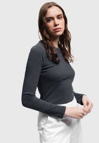 Langärmliges, geripptes Oberteil aus dunkelgrauem Stoff mit Rundhalsausschnitt und figurbetontem Schnitt, kombiniert mit weißen Hosen. Sauberes, minimalistisches Design.