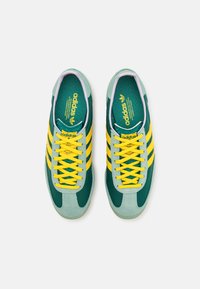 Ett par gröna Adidas-sneakers med gula ränder och snören, sedda uppifrån på vit bakgrund.