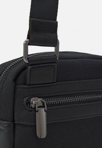 Musta laukku, jossa on teksturoitu nailonipinta. Varustettu hopeisilla yksityiskohdilla, kuten vetoketjulla ja hihnan kiinnityksellä, mikä korostaa käytännöllistä muotoilua.