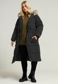 Czarna kurtka puchowa z kapturem z sztucznego futra, długa, z szerokim paskiem w talii, kieszeniami zapinanymi na zamek oraz pikowaną teksturą. Nosi się ją na zielonym swetrze z dzianiny.