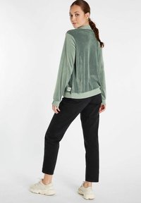 Groene, fluwelen jas met een relaxed fit en geribde mouwen, gecombineerd met zwarte broek en lichtgekleurde sneakers, wat zorgt voor een casual uitstraling.