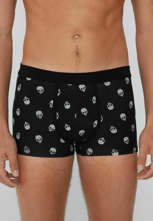 Männlicher Oberkörper in schwarzen Boxershorts mit einem grauen Totenkopf-Muster, stehend mit entspannten Armen an den Seiten vor einem schlichten Hintergrund.