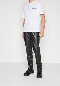 Man in een wit T-shirt met korte mouwen met tekst, zwarte neplederen broek en zwarte veterschoenen staat tegen een witte achtergrond.