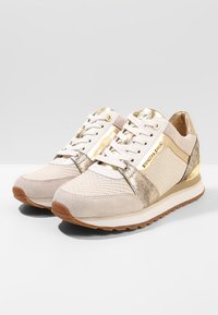 Beżowe sneakersy z teksturowaną cholewką, złotymi akcentami, białymi sznurówkami i gumową podeszwą. Charakteryzują się połączeniem materiałów, w tym zamszu i metalicznych wykończeń.