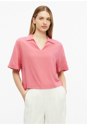 Jeune femme aux cheveux blonds et raides porte un haut rose ample à manches courtes avec col et un pantalon blanc taille haute, les mains dans les poches.