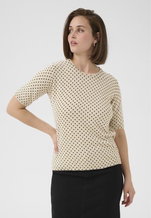 Top beige à manches courtes avec des pois noirs, col rond et bords côtelés. Associé à une jupe en jean noire. Texture douce.