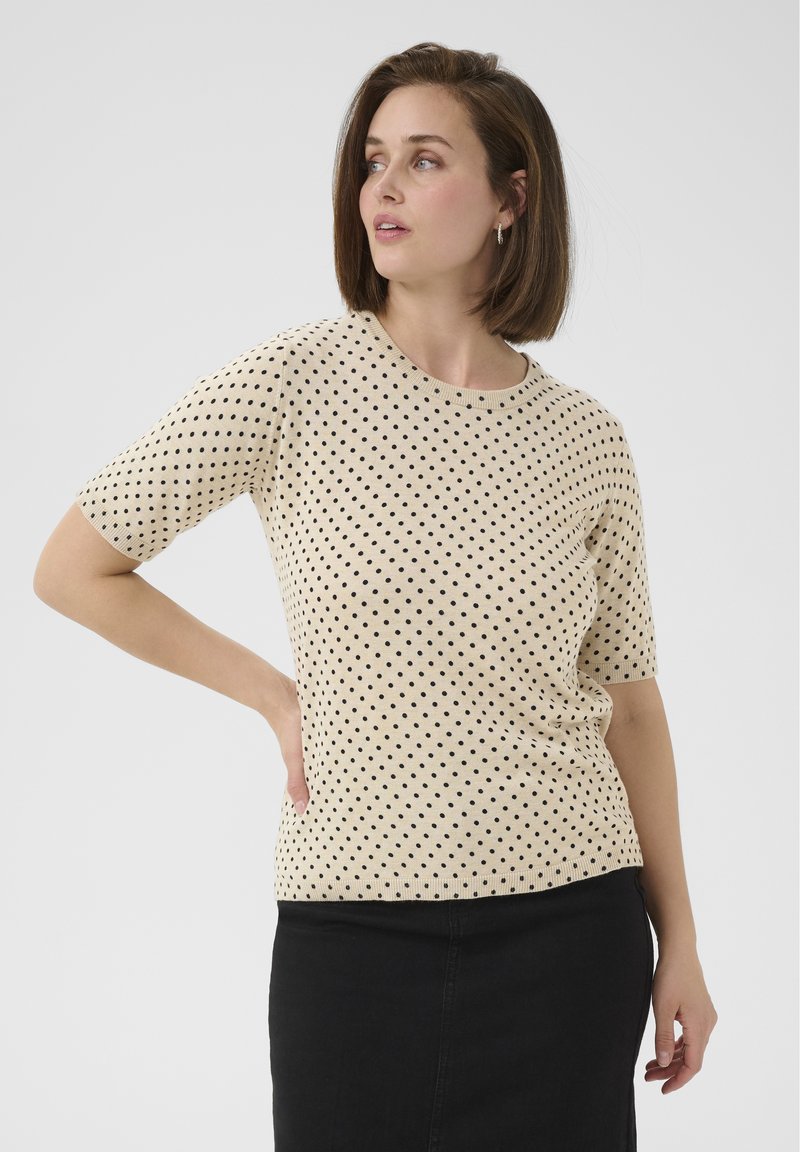 Top beige à manches courtes avec des pois noirs, col rond et bords côtelés. Associé à une jupe en jean noire. Texture douce.
