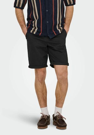 Mężczyzna w czarnych podwiniętych szortach, koszuli w paski z krótkim rękawem zapinanej na guziki, białych skarpetkach i brązowych skórzanych butach, stojący z rękami w kieszeniach.