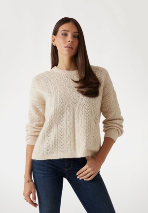 Cremfarbener gestrickter Pullover mit strukturierten Zopfmustern, gerippten Bündchen und Saum. Kombiniert mit dunkelblauen Jeans.