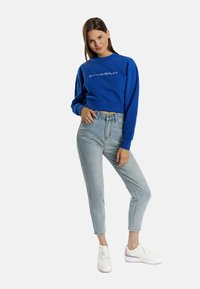 Felpa cropped blu con testo bianco e rosa, abbinata a jeans azzurri chiari e sneaker bianche con suola rosa.