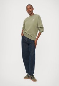 Sudadera verde oliva con cuello redondo y logo, combinada con jeans de corte suelto de color azul oscuro y zapatillas verdes con rayas negras y blancas.