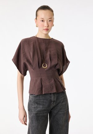 Joven mujer con blusa marrón de manga corta, vaqueros negros, aros de oro y un collar largo con un colgante redondo.