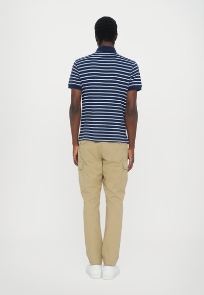 Tricou polo bleu marin cu dungi albe, mâneci scurte, asortat cu pantaloni cargo bej și adidași albi. Vedere din spate, fundal minimalist.