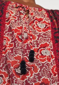 Blouse rouge à motif floral avec des détails blancs et noirs, dotée de liens à pompons noirs et d'une bordure en dentelle rouge le long des épaules.