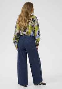 �Široke temno modre traper hlače z dodatkom okovja na straneh, v kombinaciji z večbarvno tie-dye majico. Zadnji pogled prikazuje sproščen kroj in obliko.