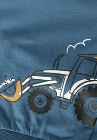 Mėlynas audinys su juoda, balta ir geltona iliustracija, vaizduojančia traktorių su priekinio kroviklio kaušu, semiančiu žemę.