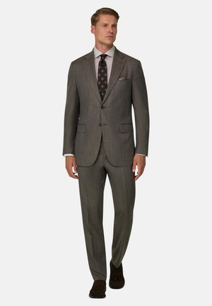 Homme portant un costume marron sur mesure avec une cravate à motifs, une chemise claire, une pochette et des mocassins marron foncé à glands, marchant vers l'avant.
