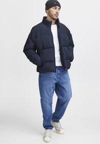 Navy puffer jacket with quilted panels, high collar, zip closure, worn over a white t-shirt, paired with blue jeans and white sneakers.