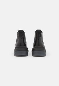 Paire de bottines en cuir noir avec semelles en caoutchouc épaisses, coutures visibles et languettes siglées à l'arrière, vues de dos.