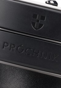 Czarny skórzany produkt z wytłoczonym logo i nazwą marki "PRÓCHNIK", z błyszczącymi elementami metalowymi i teksturowaną powierzchnią.