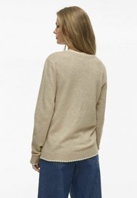 Suéter de punto beige con cuello redondo, mangas largas y delicado ribete de pompones en el bajo. Combinado con jeans de mezclilla azules.