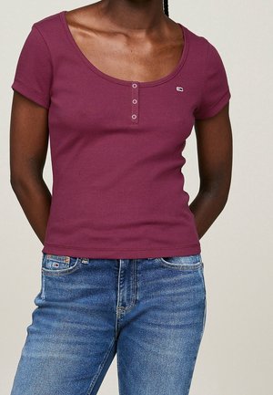 Femme portant un haut henley bordeaux à manches courtes ajusté avec des détails à boutons, associée à un jean en denim bleu, debout devant un fond uni.