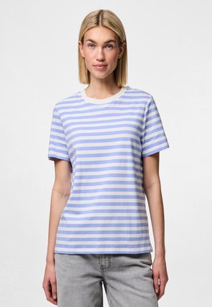 Femme blonde debout face à l’avant, portant un t-shirt à manches courtes rayé horizontalement bleu clair et blanc et un pantalon gris clair.