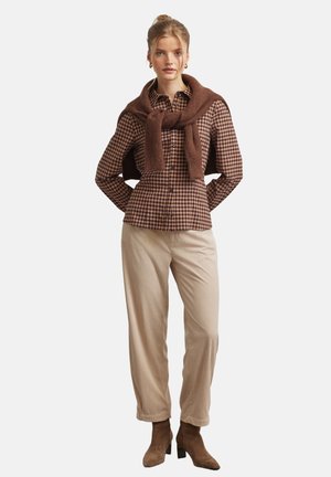 Frau steht und trägt ein braun kariertes Hemd, beige Hose, braune Knöchelstiefel und einen braunen Pullover, der über ihre Schultern gelegt ist.