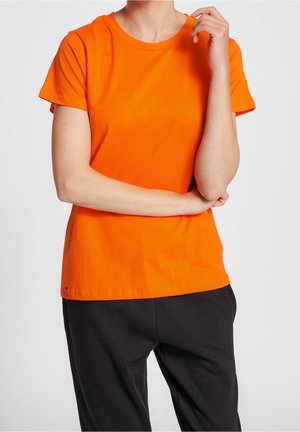 Persoon in een oranje T-shirt met korte mouwen en een zwarte broek, staand met één arm over de borst en de andere hand nabij het gezicht.