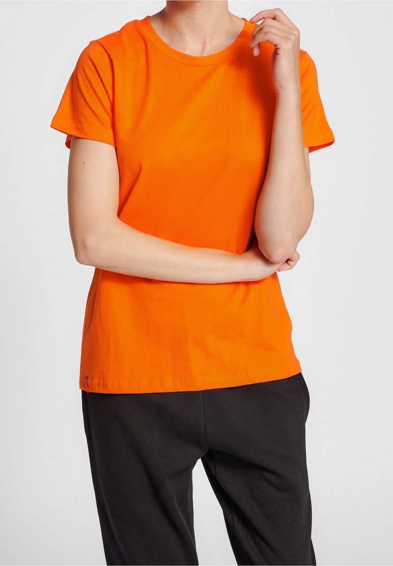 Personne portant un t-shirt orange à manches courtes et un pantalon noir, debout avec un bras croisé et l'autre main près du visage.