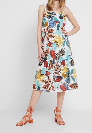 Vrouw draagt een mouwloze jurk tot op de knie met kleurrijke tropische bladpatronen en oranje sandalen met blokhak en bandjes.