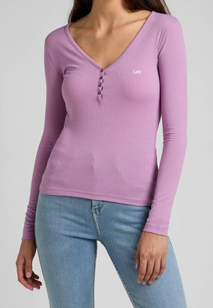 Lange mouwen, ribbelige paarse top met V-hals, met vijf knoopdetails en een klein logo aan de linkerkant. Zachte stoftextuur.