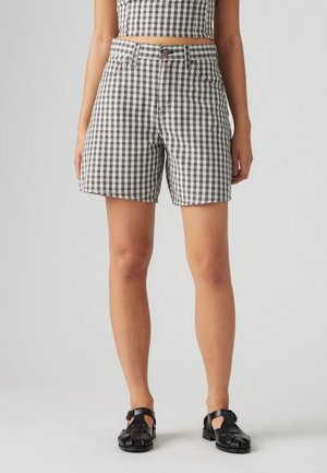 Kvinde iført sorte og hvide gingham højttaljede shorts, matchende cropped top og sorte lukkede sandaler, stående på en ensfarvet grå baggrund.