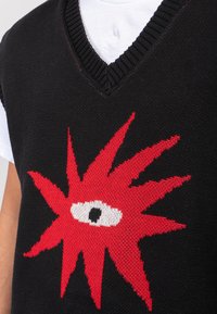 Μαύρο πλεκτό γιλέκο με σχέδιο σε σχήμα V που εμφανίζει ένα έντονο κόκκινο μοτίβο αστραπής και μια κεντρική λευκή λεπτομέρεια ματιού.