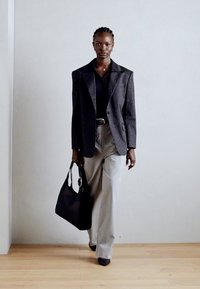 Blazer noir à rayures sur un top noir, pantalon large gris clair, et un sac noir. Textures lisses avec des épaules prononcées et une quincaillerie circulaire.
