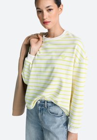 Weißer Pullover mit gelben Streifen, locker geschnitten, mit gerippten Bündchen und einem kleinen bestickten Logo. Dazu hellblaue Jeans aus Denim.