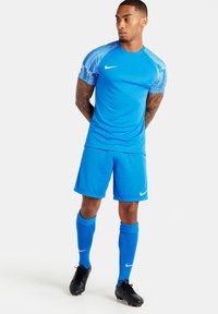 Blaues Nike-Sportoutfit mit kurzen Ärmeln, gestreiftem Schulterdesign, passenden Shorts, knielangen Socken und schwarzen Fußballschuhen.