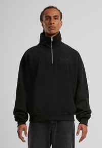 Svart zip-up hettegenser laget av myk materiale, med høy krage og ribbestrikkede mansjetter, med subtil logo-detaljering på brystet.