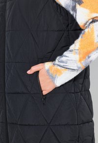 Svart quiltad väst med sidodragkedja och en hand i fickan. Vesten har en jämn, matt textur med diamantsydda stygn.