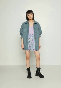 Mulher asiática em pé, vestindo um casaco oversized teal, uma blusa lavanda, uma saia azul com flores e botas de combate pretas com atacadores, contra um fundo simples.