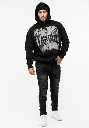 Czarny sweatshirt z dużą szaro-srebrną grafiką "TAPOUT", zestawiony z zniszczonymi czarnymi dżinsami i białymi sneakersami. Model nosi kaptur.