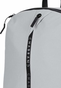 Sac à dos gris clair en tissu avec une finition texturée. Dispose d'une fermeture éclair noire et d'un panneau latéral vertical noir avec le logo.