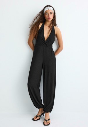 Donna con capelli ricci che indossa una tuta nera senza maniche e una fascia bianca a pois, in piedi contro una parete chiara e semplice.