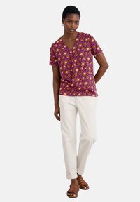 Top cu mânecă scurtă, de culoare mov, cu decolteu în V și motive florale în nuanțe de roz și portocaliu, asociat cu pantaloni albi și sandale maro cu barete.