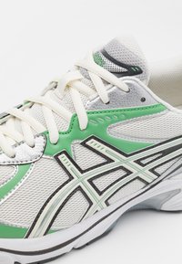 Zapatilla deportiva multicolor con parte superior de malla blanca y acentos en verde y plata, detalles negros y cordones texturizados. Suela de goma.