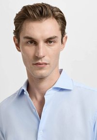 Modra karo srajca s ovratnikom, nežno teksturo in sprednjim zapenjanjem na gumbe. Model ima kratke lase in nevtralen izraz.