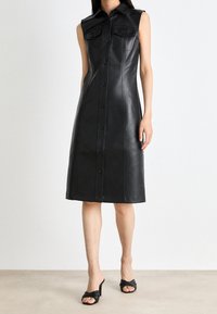Zwarte mouwloze leren jurk met een knoopsluiting aan de voorkant, twee borstzakken en een knielange A-lijn met zichtbare naden.