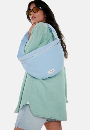 Femme portant des lunettes de soleil à verres jaunes, une chemise oversize vert clair, un short en jean, et portant un grand sac à bandoulière bleu clair avec fermeture éclair visible et étiquette.