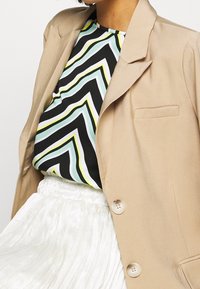 Beige blazer med en enkelt lomme og to knapper, parret med en top med zigzag-mønster i sort, grøn og hvid. Tekstureret hvid nederdel nedenunder.