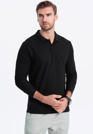 Camisa polo negra de manga larga en algodón con cuello clásico, tapeta de dos botones y textura suave. Llevada por un hombre con un reloj.